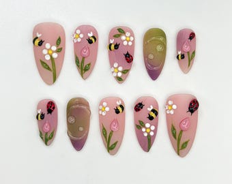 Puede incluir: Un conjunto de diez uñas postizas en forma de almendra. Cada uña está pintada de color rosa claro con diseños florales e insectos. Los diseños incluyen flores blancas, hojas verdes, abejas amarillas y negras y mariquitas rojas.