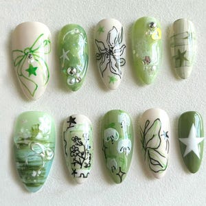 Puede incluir: Diez uñas postizas ovaladas de color verde y blanco con diferentes diseños. Los diseños incluyen un lazo, flores, estrellas y una mariposa.