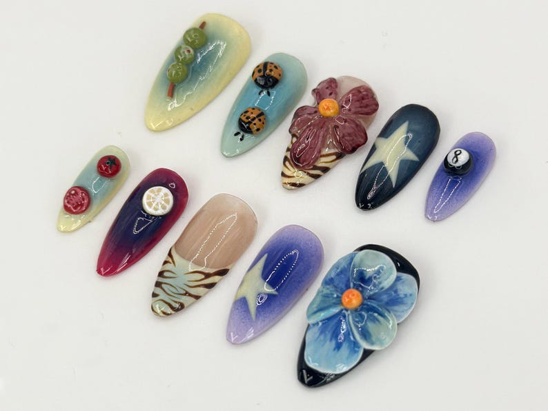 K&ouml;nnte beinhalten: Zehn verschiedene Nageldesigns mit verschiedenen Themen, darunter ein Marienk&auml;fer, eine Blume, eine Zitrone, ein Stern und eine 8-Ball. Die N&auml;gel sind in Blaut&ouml;nen, Lila, Rosa und Gelb lackiert.