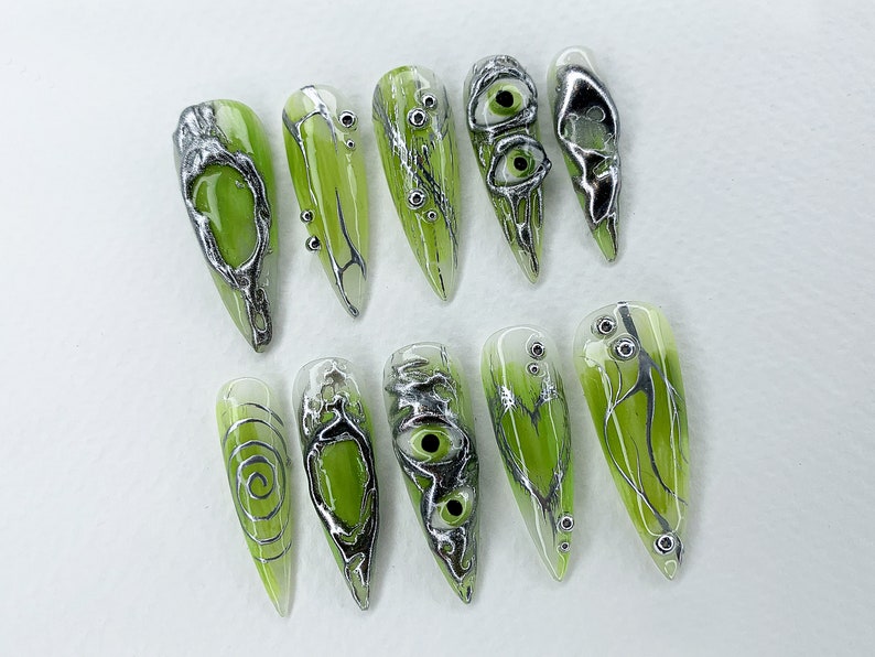 May include: Set of ten green and silver nail tips with various designs, including swirls, eyes, and branches.