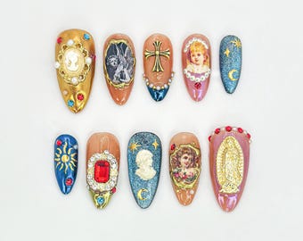 May include: Ten long, almond-shaped artificial nails with various designs. The designs include a gold cross, a sun, a crescent moon, a red gemstone, a cameo, and religious figures.