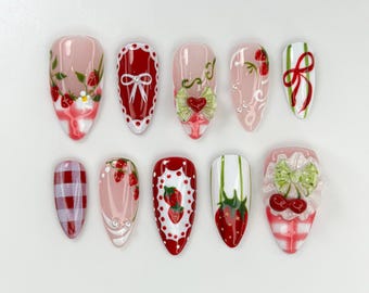 May include: A set of ten almond-shaped false nails with various designs. The designs include strawberries, bows, and gingham patterns in red, white, and pink. Some nails feature 3D embellishments.