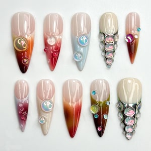 May include: Ten long, almond-shaped fake nails with different ombre designs and embellishments. Some nails have iridescent gems, others have small pearls, and some have a silver accent.