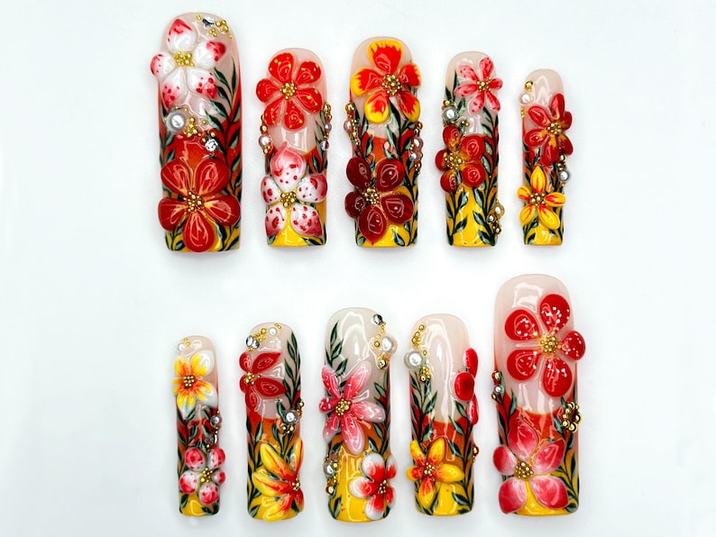 May include: A set of ten artificial nails, each adorned with 3D floral designs. The nails feature a base of yellow and white, with red, white, and yellow flowers, green leaves, and gold accents. The nails are arranged on a white surface.