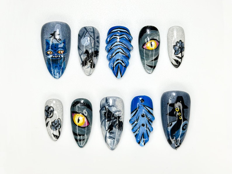 May include: A set of ten artificial nails with various designs. The nails feature a mix of blue, gray, and silver colors. Designs include a cat, an eye, and abstract patterns. The nails are oval-shaped and are arranged on a white surface.