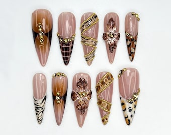 May include: A set of ten long, almond-shaped artificial nails. The nails feature various designs, including animal prints, gold embellishments, and abstract patterns in shades of brown, beige, and gold. The nails are arranged on a white surface.