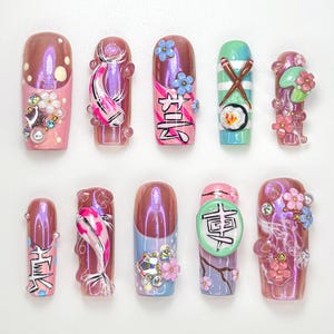 May include: A set of ten artificial nails with various designs. The nails feature a range of colors, including pink, purple, and green, and are adorned with embellishments such as flowers, koi fish, and Japanese characters. Some nails have a glossy finish.