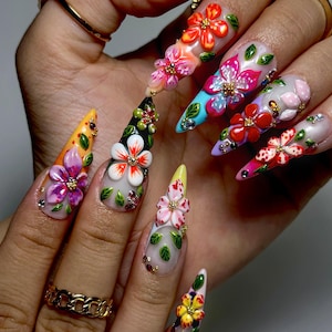 May include: A hand showing long, pointed acrylic nails, each decorated with 3D floral designs. The nails feature various colours, including orange, purple, white, and pink, with intricate details like rhinestones and gold accents. The overall theme is a vibrant, artistic nail design.
