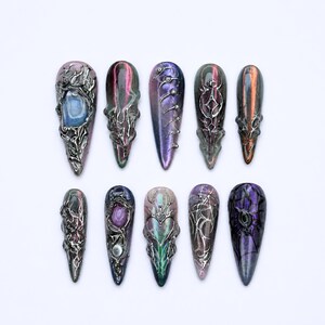 May include: A set of ten almond-shaped press-on nails featuring a dark, iridescent base with metallic silver 3D relief designs. The patterns include intricate swirling lines, raised spikes, and embedded circular accents in shades of purple, blue, and copper.