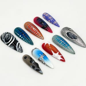 May include: Ten long, almond-shaped false nails with various designs. The designs include black and white swirls, blue and white dots, red and white stripes, a silver and blue geometric pattern, a red and white ombre, a blue and white ombre, a silver and blue geometric pattern, a silver and black design, a silver and blue geometric pattern, and a silver and black design.