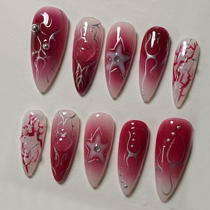 May include: Set of ten long, stiletto-shaped false nails with a gradient design. The nails are painted with a white to red gradient and feature silver glitter, hearts, and stars.