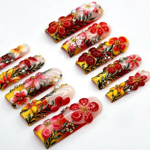 May include: A collection of press-on nails with intricate floral designs. Each nail features a base of yellow, red, and white, adorned with detailed flower art, gold accents, and small embellishments. The nails are arranged on a white surface.