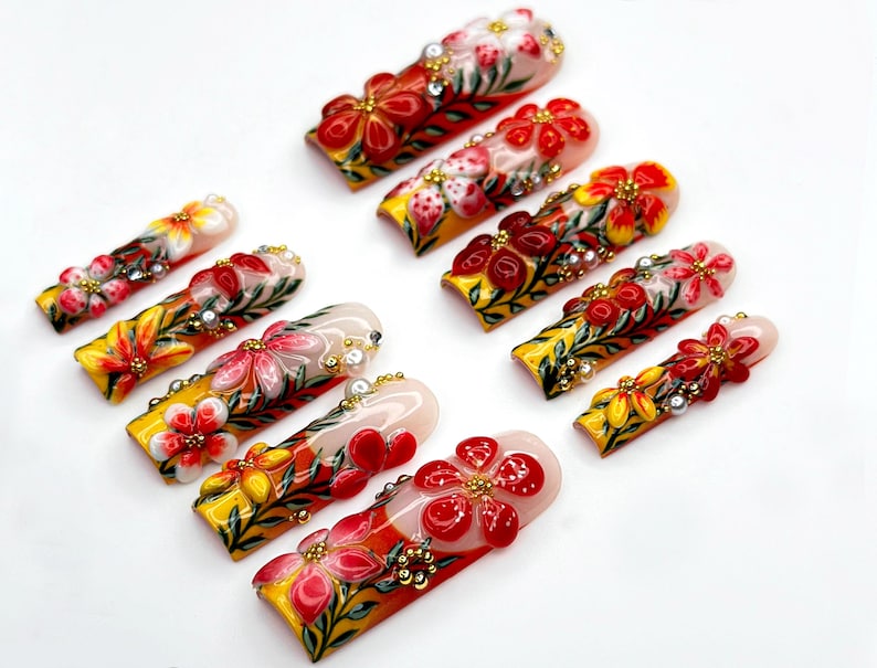 May include: A collection of press-on nails with intricate floral designs. Each nail features a base of yellow, red, and white, adorned with detailed flower art, gold accents, and small embellishments. The nails are arranged on a white surface.