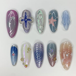 May include: Ten different nail designs featuring a variety of colors, patterns, and embellishments. The designs include a pink nail with silver stars, a blue nail with a silver star, a white nail with silver swirls, and a pink nail with a silver star.