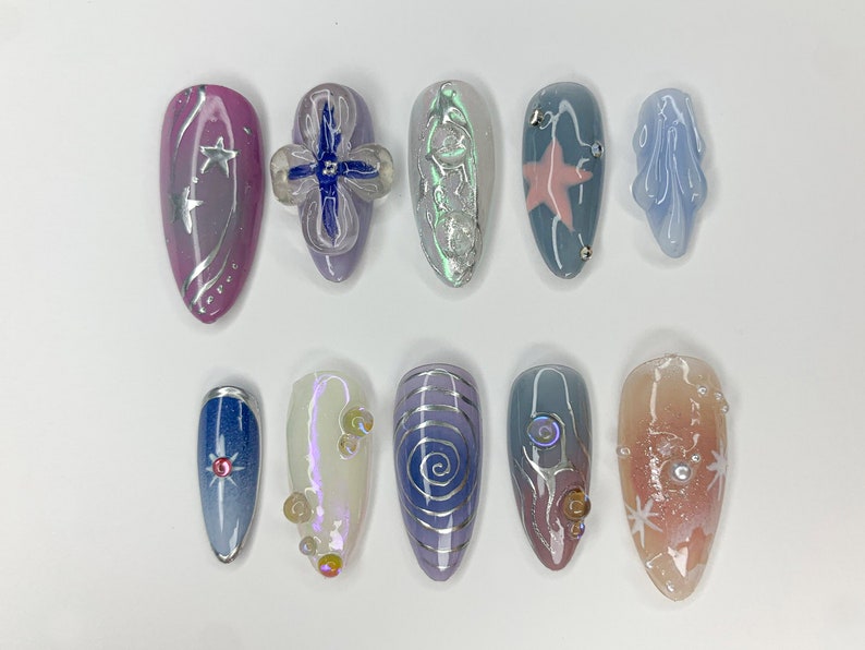 May include: Ten different nail designs featuring a variety of colors, patterns, and embellishments. The designs include a pink nail with silver stars, a blue nail with a silver star, a white nail with silver swirls, and a pink nail with a silver star.