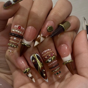 May include: A hand displaying long, pointed acrylic nails. The nails feature a variety of designs, including brown, gold, and white accents, with embellishments like pearls, gems, and metallic rings. Some nails have a French tip design.