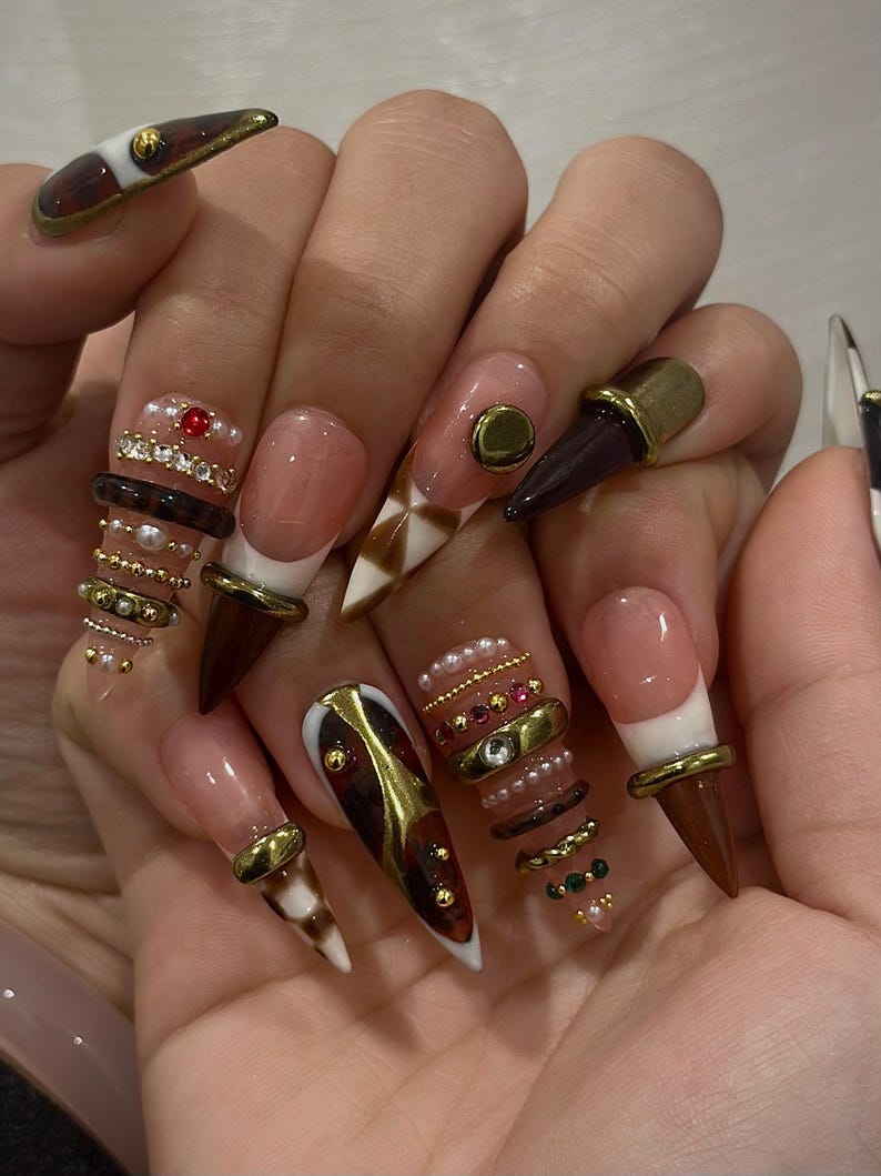 May include: A hand displaying long, pointed acrylic nails. The nails feature a variety of designs, including brown, gold, and white accents, with embellishments like pearls, gems, and metallic rings. Some nails have a French tip design.