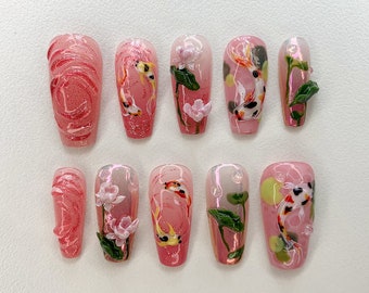 Puede incluir: Diez uñas postizas rosas y blancas con un diseño de peces koi y flores. Las uñas son largas y de forma ovalada.