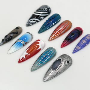 May include: A collection of ten long, almond-shaped false nails with various designs. The nails are painted in a variety of colors, including black, white, blue, red, and purple. Some of the nails have intricate designs, such as swirls, lines, and geometric patterns.