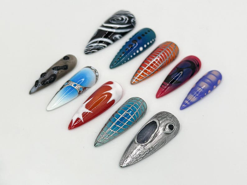 May include: A collection of ten long, almond-shaped false nails with various designs. The nails are painted in a variety of colors, including black, white, blue, red, and purple. Some of the nails have intricate designs, such as swirls, lines, and geometric patterns.
