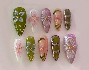 Könnte beinhalten: Eine Sammlung von zehn künstlichen Nägeln, jeweils mit einem einzigartigen Design. Die Nägel weisen verschiedene Farben auf, darunter Grün, Rosa und Lila, und sind mit floralen Verzierungen und goldenen Akzenten verziert. Einige Nägel haben ein glitzerndes Finish.