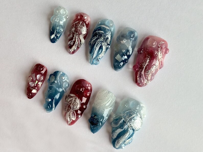 May include: Set of ten false nails with a gradient design in shades of blue, red, and white. Each nail features a silver glitter design and a 3D embellishment.