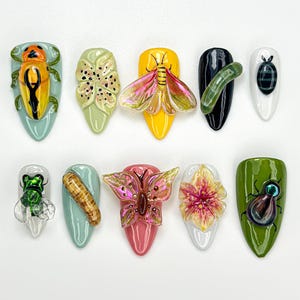 Pode incluir: Uma coleção de dez peças de nail art com designs intrincados. Cada unha apresenta um design diferente de inseto ou floral, incluindo besouros, borboletas e flores, em cores vibrantes como amarelo, verde, rosa e preto. As unhas são em formato amêndoa e sobre um fundo branco.
