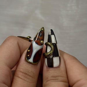 May include: Two long, almond-shaped fingernails with unique designs. One nail features a brown, white, and gold design with gold accents. The other nail has a black and white checkerboard pattern with a gold crescent detail. Nail art.