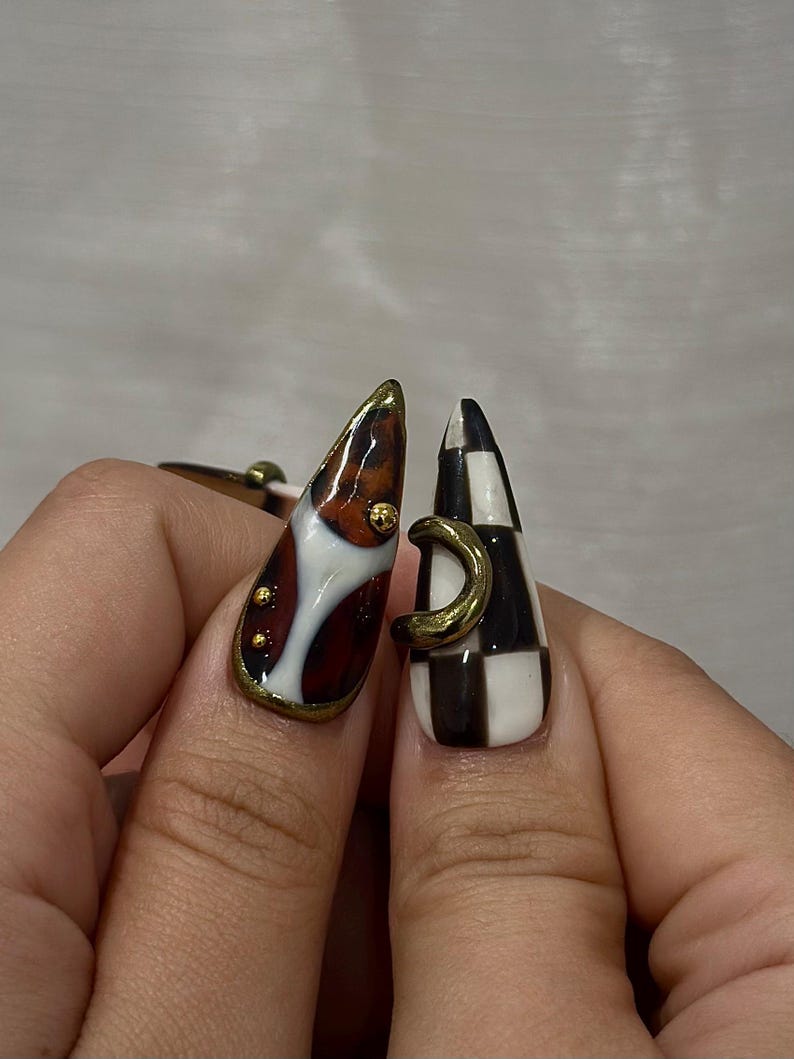 May include: Two long, almond-shaped fingernails with unique designs. One nail features a brown, white, and gold design with gold accents. The other nail has a black and white checkerboard pattern with a gold crescent detail. Nail art.