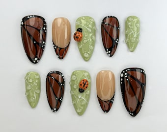 May include: Set of ten false nails featuring a butterfly and ladybug design. The nails are in a range of colours, including brown, green, and white.