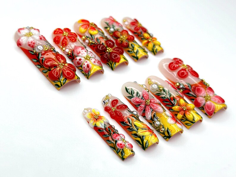 May include: A set of press-on nails with a floral design. The nails feature a variety of colors, including red, yellow, and white, with intricate flower details and embellishments. The nails are arranged on a white surface.