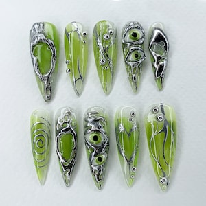 Puede incluir: Diez diseños de arte de uñas verdes y plateados con remolinos, ojos y ramas. Los diseños están sobre un fondo blanco.