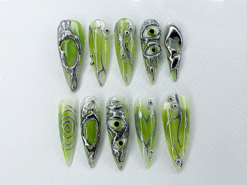 May include: Ten green and silver nail art designs featuring swirls, eyes, and branches. The designs are on a white background.