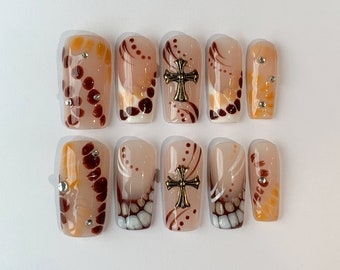May include: Set of ten false nails with a brown, orange, and white abstract design. Some nails feature a gold cross accent.
