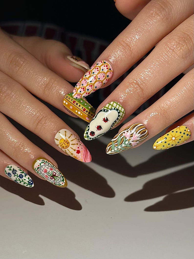 May include: A set of long, almond-shaped fingernails, each painted with a unique design. The designs include floral patterns, suns, and geometric shapes in a variety of colors, including pink, yellow, green, and white. The nails are well-manicured and showcase intricate nail art.