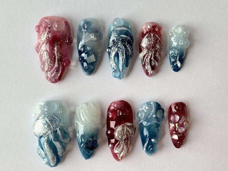 May include: Set of ten false nails with a gradient design. The nails are decorated with white and silver swirls, and some have small pearl-like embellishments. The nails are in two color schemes: blue and white, and red and white.