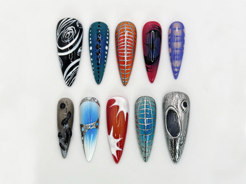 May include: A set of 12 long, almond-shaped false nails with various designs. The designs include black and white swirls, teal with white dots, orange with silver lines, red with black swirls, purple with white dots, gray with black swirls, blue with silver accents, red with white swirls, teal with silver lines, silver with a black eye, and a silver nail with a black eye.