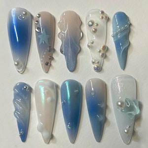 May include: Set of ten long, oval-shaped artificial nails with a variety of blue and white ombre designs. Some nails feature small, iridescent beads, a star, or a swirl pattern.