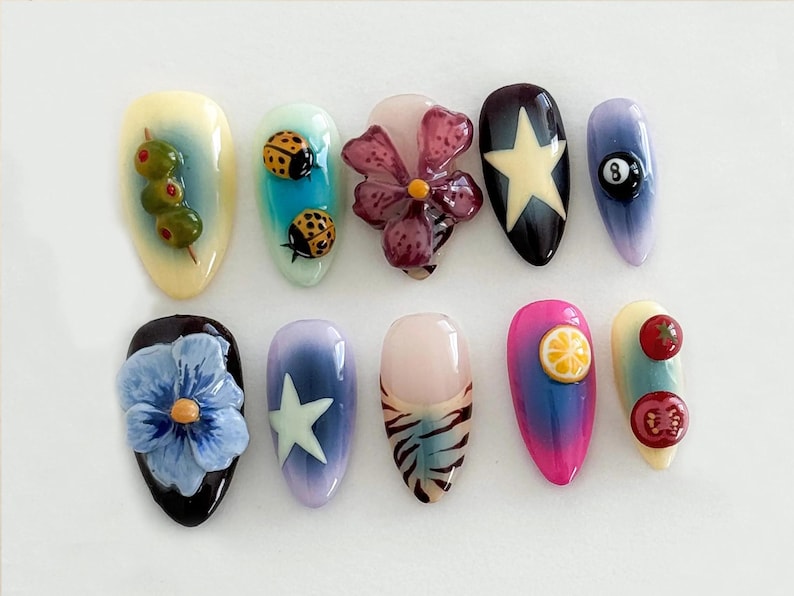 May include: Ten different nail designs featuring various themes, including ladybugs, olives, flowers, a star, a pool ball, a lemon, and tomatoes.