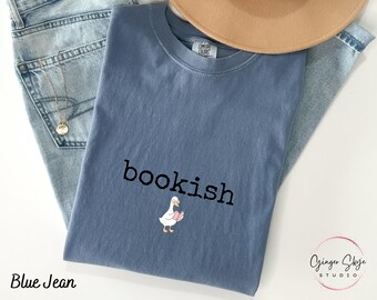 Puede incluir: Una camiseta azul vaquera con la palabra "bookish" impresa en negro, junto con un ganso de dibujos animados leyendo un libro. La camiseta está doblada sobre unos vaqueros azules y un sombrero beige.