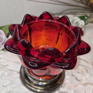 May include: A vibrant red glass candle holder with a flower-like design. The candle holder has a silver-coloured base and a transparent appearance. The petals of the flower are pointed and create a unique shape. The candle holder is a decorative item.