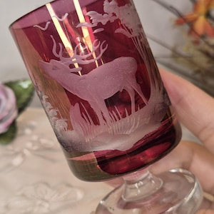 May include: A burgundy glass with a clear base, showcasing an etched design of a stag in a woodland setting. The glass has a short stem and a wide, slightly flared rim. The stag is the central element of the design.