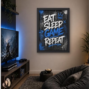 Könnte beinhalten: Ein gerahmter Kunstdruck mit dem Schriftzug "EAT SLEEP GAME REPEAT" in weißer und blauer Graffiti-Schrift auf schwarzem Hintergrund. Der Druck befindet sich in einem schwarzen Rahmen und wird in einem Wohnzimmer gezeigt.