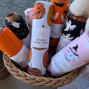 May include: A wicker basket holds several rolls of reusable flannel towels. The towels feature Halloween-themed patterns, including jack-o'-lanterns, bats, ghosts, and a witch cat. Each roll is labeled "REUSABLE FLANNEL TOWELS 12 PACK".