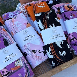 May include: A display of reusable flannel towels with Halloween-themed patterns. The towels are bundled and wrapped with white labels that read "REUSABLE FLANNEL TOWELS". The patterns include ghosts, pumpkins, cats, and bats in colors like purple, orange, pink, and black.