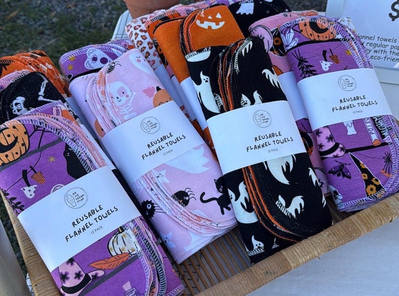 May include: A display of reusable flannel towels with Halloween-themed patterns. The towels are bundled and wrapped with white labels that read "REUSABLE FLANNEL TOWELS". The patterns include ghosts, pumpkins, cats, and bats in colors like purple, orange, pink, and black.