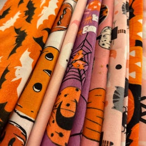 May include: A stack of Halloween-themed fabric rolls in various colors. The fabrics feature orange, pink, and purple backgrounds with Halloween-related patterns, including bats, pumpkins, spiderwebs, and cats. The fabric rolls are tightly packed together.