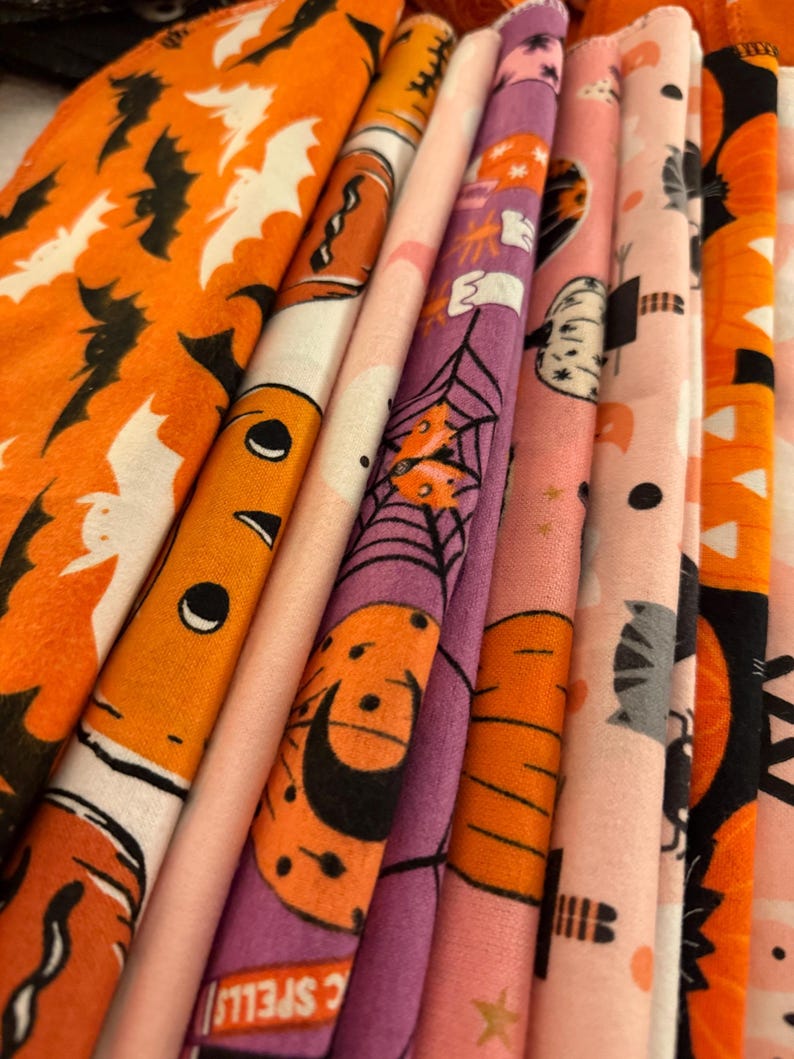 May include: A stack of Halloween-themed fabric rolls in various colors. The fabrics feature orange, pink, and purple backgrounds with Halloween-related patterns, including bats, pumpkins, spiderwebs, and cats. The fabric rolls are tightly packed together.