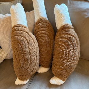 May include: Three plush turkey leg pillows with brown and white textured fabric. The pillows are arranged on a brown couch with other pillows in the background. The turkey leg pillows have a realistic design.