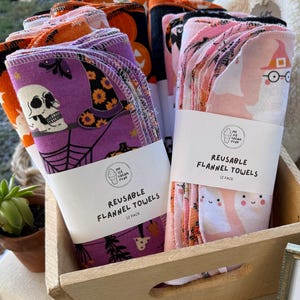 May include: Stacks of reusable flannel towels in a wooden crate. The towels feature Halloween-themed designs, including skulls, witch hats, pumpkins, and ghosts. Each pack is labeled "REUSABLE FLANNEL TOWELS 12 PACK".